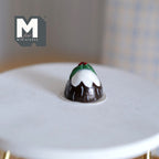 1:12 scale miniature Christmas pudding with white icing and holly on top, shown on a white round table