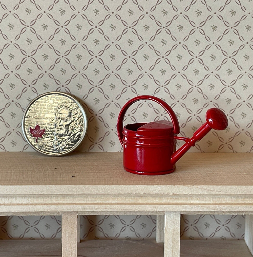1:12 scale miniature metal watering can in red with spray nozzle, shown on a wood shelf beside a coin for scale