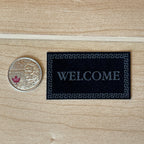 1:12 scale miniature felt welcome door mat in black with a gray Greek key border, shown on a wood floor beside a coin