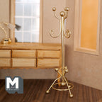 1:12 scale miniature gold metal coat rack with curled hooks and round base, standing in a room beside a wood desk