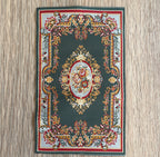 1:12 scale miniature woven area rug in green with floral medallion and red and gold border, on a wood floor