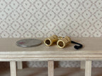 1:12 scale miniature metal binoculars in brass with black strap, shown on a wood shelf beside a coin for scale