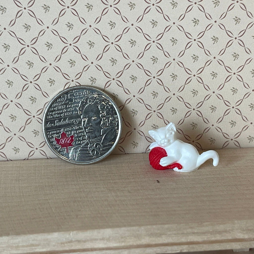 1:12 scale miniature white kitten playing with a red yarn ball, shown on a wood shelf beside a coin for scale