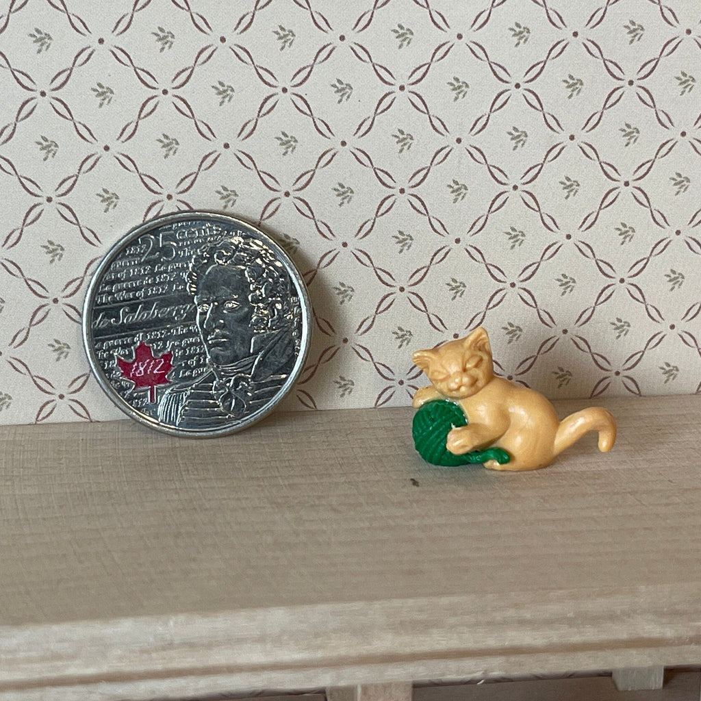 1:12 scale miniature orange kitten playing with a green yarn ball, on a wood shelf beside a coin for scale