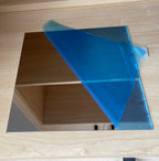 1:12 scale miniature flexible mirror sheet with blue protection film partly peeled back, shown on a wood surface