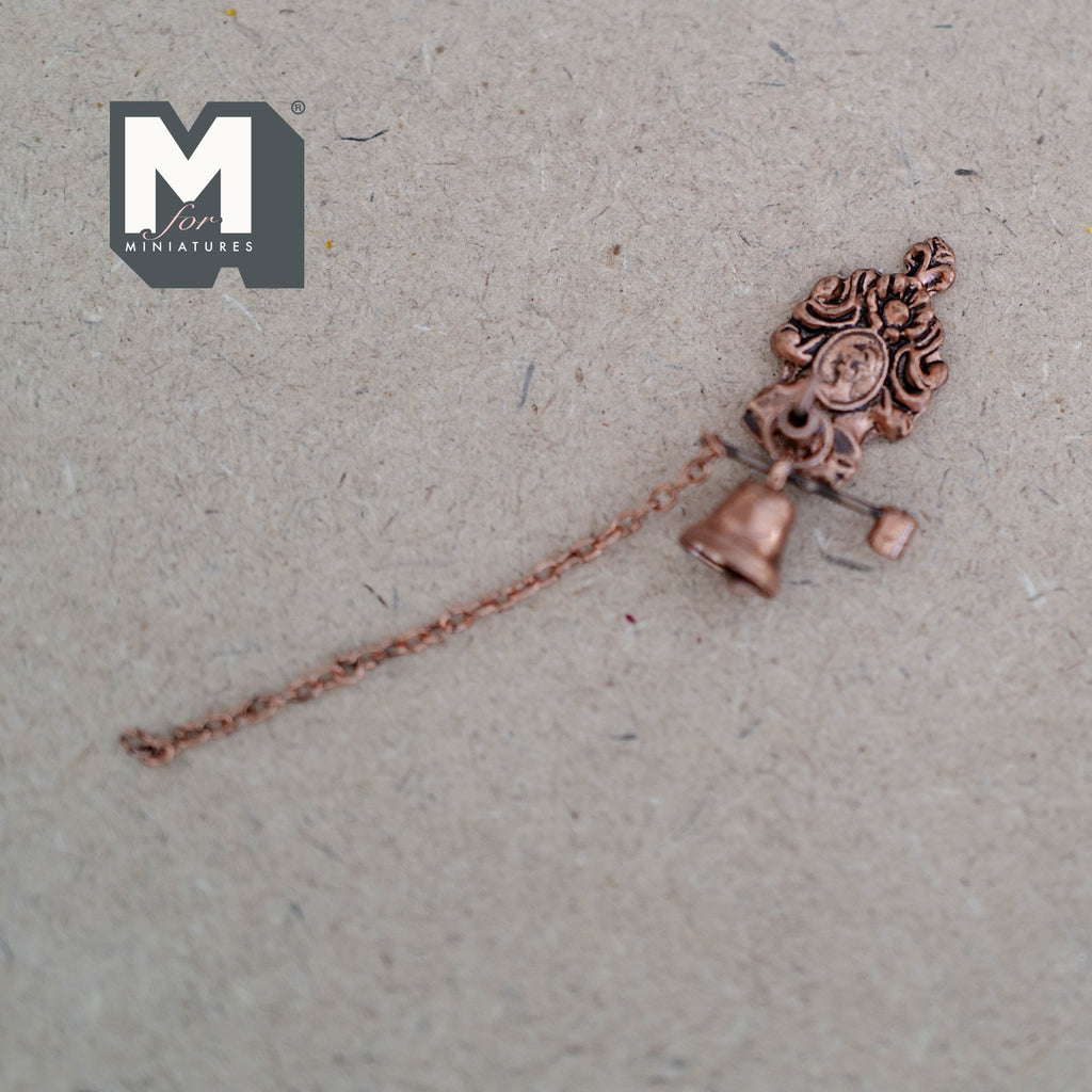 1:12 scale miniature metal house bell in bronze with scroll bracket and pull chain, shown laid on a tan surface