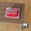 1:12 scale miniature clay American flag cake with red and white stripes on a white doily, shown on a wood table
