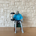 1:12 scale miniature black dumbbell with 4 removable plates, lifted by a seated artist figure by a stone wall