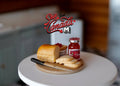 1:12 scale miniature breakfast board with sliced bread loaf, knife and jam jar, shown on a white round table