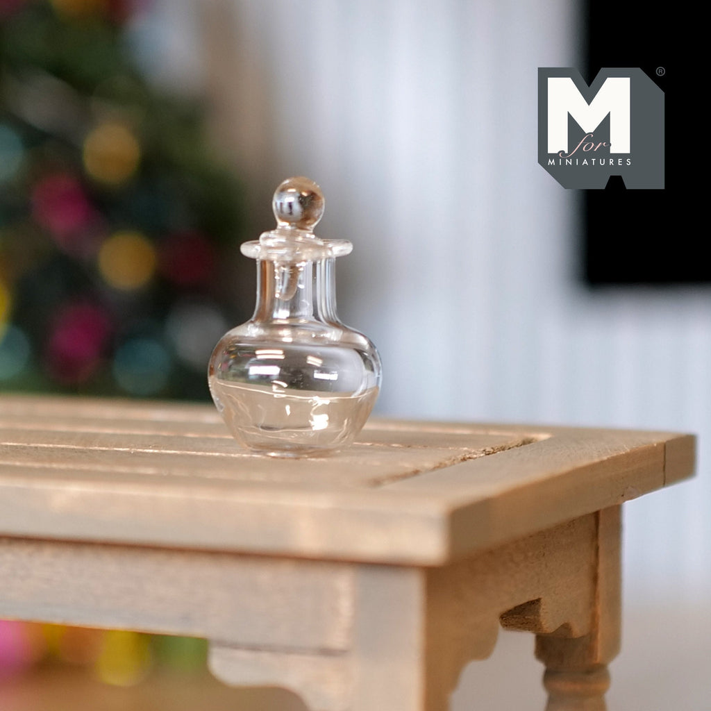 1:12 scale miniature clear glass wine decanter with stopper, round body, shown on a wood table with holiday lights behind