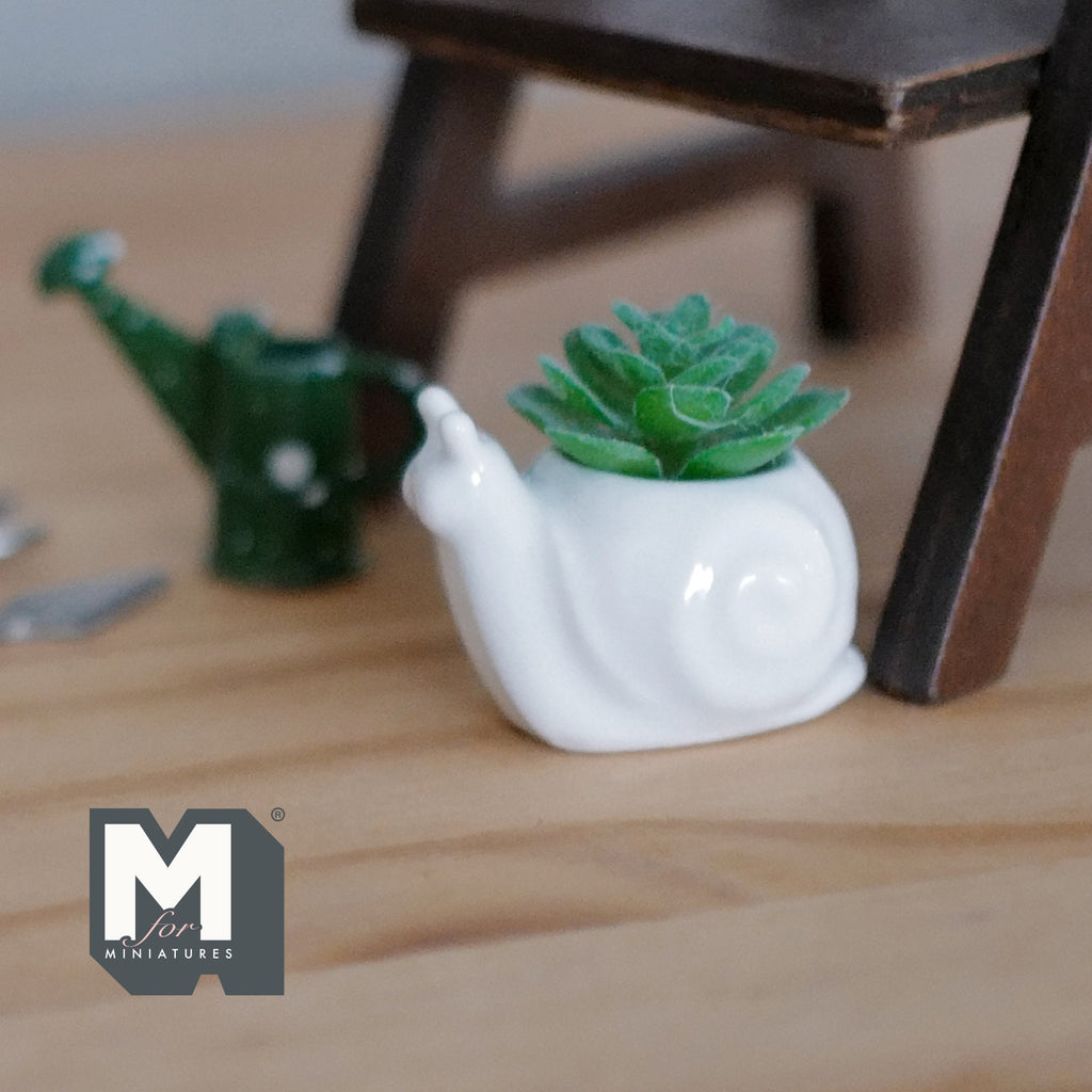1:12 scale miniature ceramic snail planter in white with green succulent, shown on a wood floor by a watering can