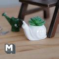 1:12 scale miniature ceramic snail planter in white with green succulent, shown on a wood floor by a watering can