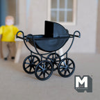 1:12 scale miniature metal baby pram in black with spoked wheels, curled handle and folding hood, shown outside a house