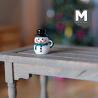 1:12 scale miniature Christmas snowman mug with lid in white with a black top hat and green buttons, shown on a wood table