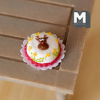 1:12 scale miniature reindeer Christmas cake with white frosting and yellow flowers on a doily, shown on a wood table