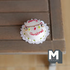 1:12 scale miniature clay Merry Christmas cake with white icing, red lettering and garland on a doily, on a wood table