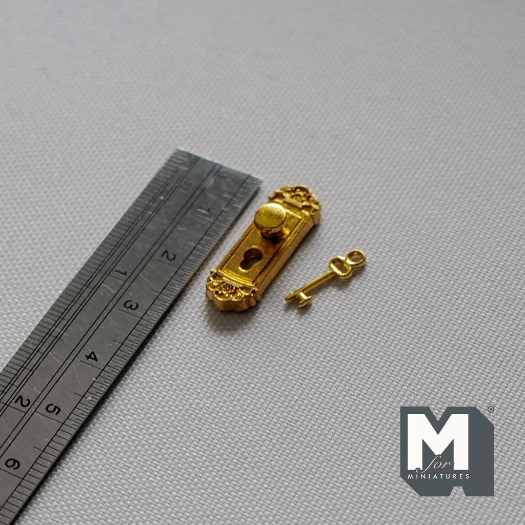 1:12 scale miniature gold door knob plate with key, ornate backplate shown beside a metal ruler on gray fabric