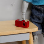 1:12 scale miniature metal tape dispenser in red with real tape, shown on a wood desk beside a jointed figure