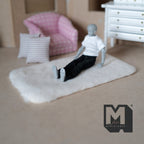 1:12 scale miniature cream faux sheepskin rug, rectangular with rounded corners, shown on a floor with a seated figure