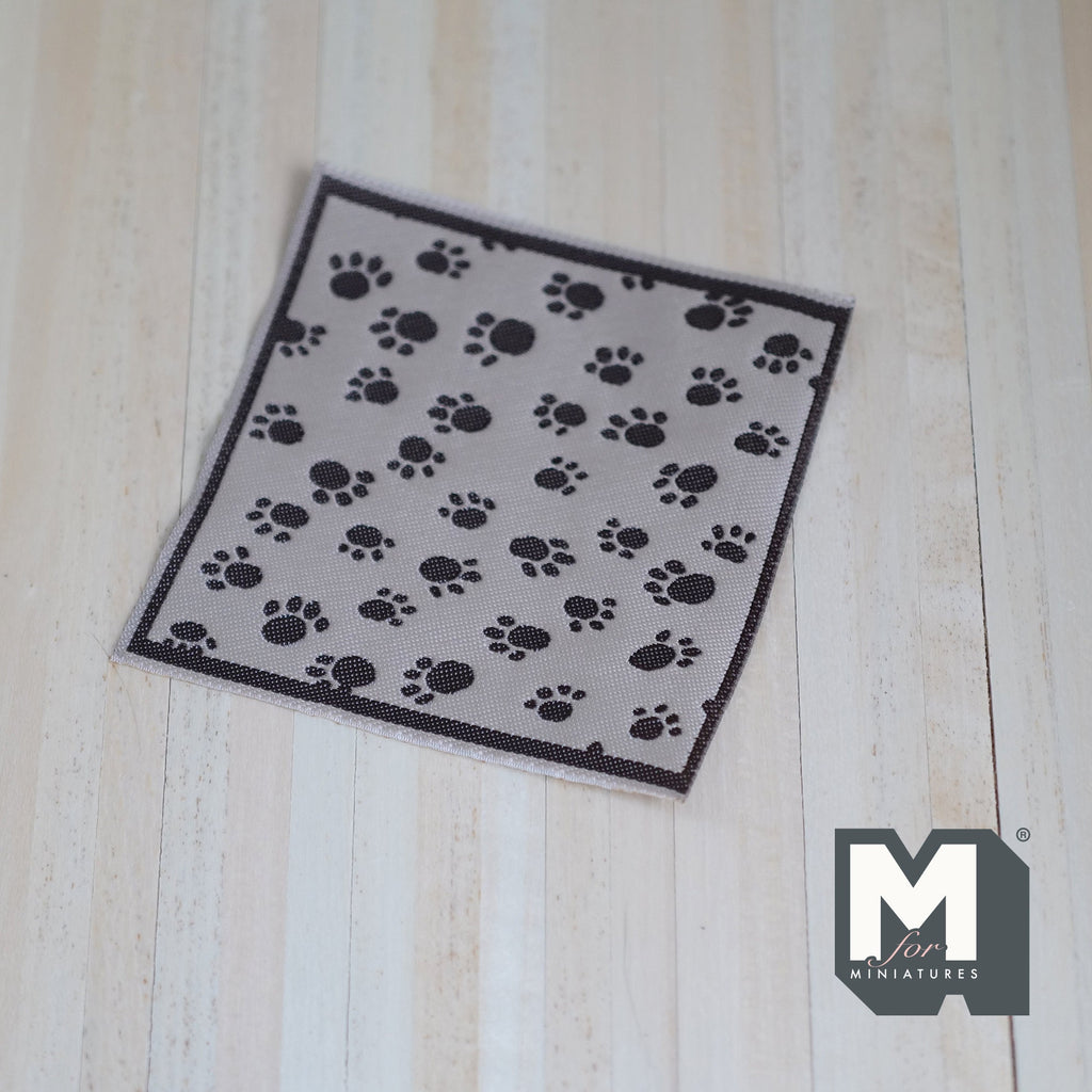 1:12 scale miniature pet blanket in beige with black paw print pattern and dark border, on a wood floor
