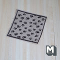 1:12 scale miniature pet blanket in beige with black paw print pattern and dark border, on a wood floor