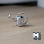 1:12 scale miniature metal watering can in silver with spray nozzle and loop handle, shown on a wood floor