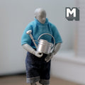 1:12 scale miniature metal watering can in silver with spray nozzle and handle, shown held by a poseable figure