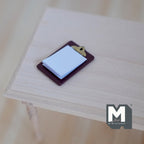 1:12 scale miniature clipboard in burgundy with gold clip and white note pad, shown on a light wood table