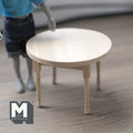 1:12 scale miniature unpainted wood round dining table with turned legs, shown beside a poseable figure for scale