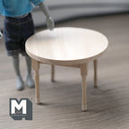 1:12 scale miniature unpainted wood round dining table with turned legs, shown beside a poseable figure for scale