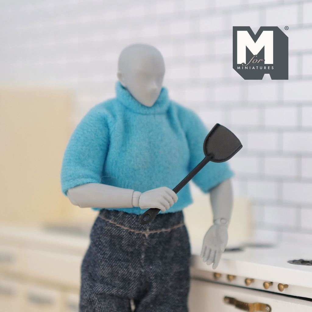 1:12 scale miniature metal slotted turner in black with long handle, held by an artist figure in a white kitchen