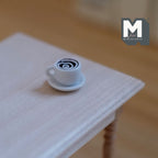 1:12 scale miniature cup of hot chocolate with swirl top on a white saucer, shown on a white wood table