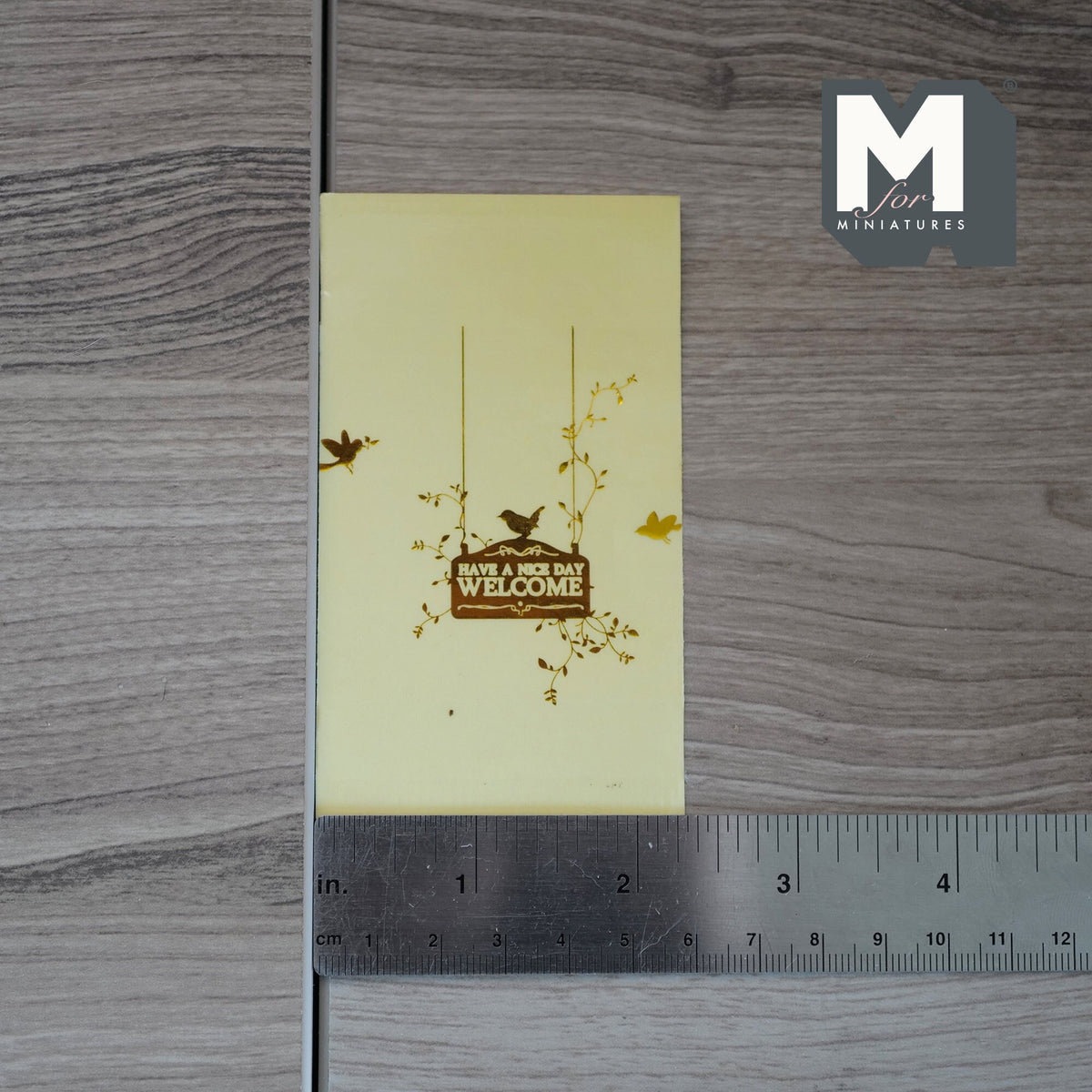 1:12 scale miniature gold welcome have a nice day decal with birds and leaves, shown on backing sheet with a ruler