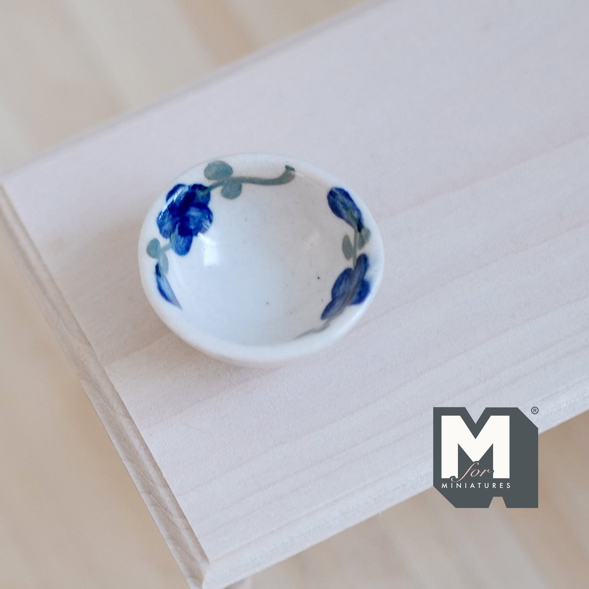 1:12 scale miniature ceramic bowl in white with dark blue floral rim, shown on a light wood tabletop