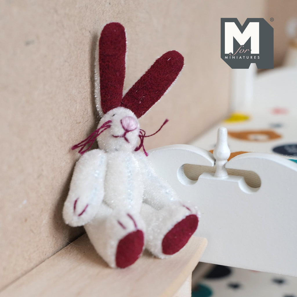 1:12 scale miniature white plush lop bunny rabbit with movable arms and legs, dark red ears and feet, sitting by a bed