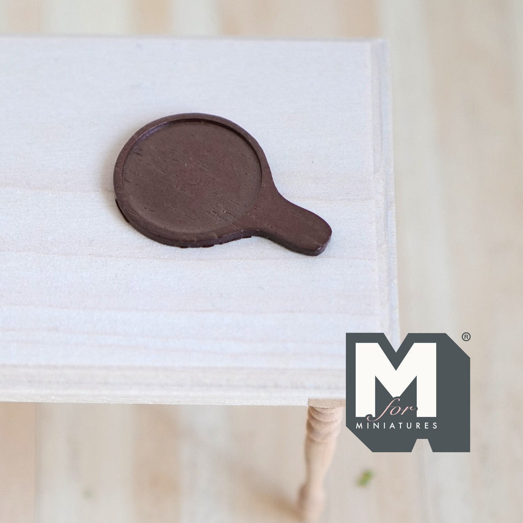 1:12 scale miniature wood round cheese board with handle in dark brown, shown lying on a white wood table