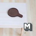 1:12 scale miniature wood round cheese board with handle in dark brown, shown lying on a white wood table