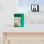 1:12 scale miniature metal tea tin with lid in green with a printed label, shown on a bare wood table