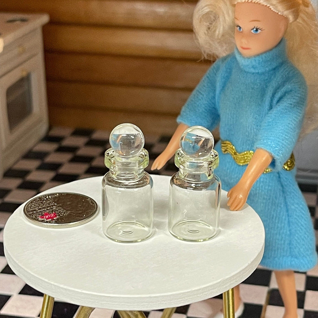 1:12 scale miniature clear glass canisters with glass lids, set of 2, shown on a white table beside a coin and a doll
