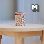 1:12 scale miniature metal tea tin with lid in cream with floral print, shown on a round wood table