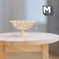 1:12 scale miniature large cream metal wire fruit basket with open loop rim and pedestal foot, on a round wood table