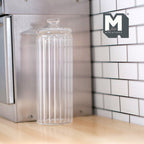 1:12 scale miniature clear ribbed glass canister with lid, tall cylinder, shown on a kitchen counter by white tile