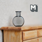 1:12 scale miniature gray glass bottle vase with flat round body and short neck, shown on a wood dresser