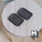 1:12 scale miniature ceramic rectangular plates in dark brown with raised rims, set of 2, shown on a round wood table