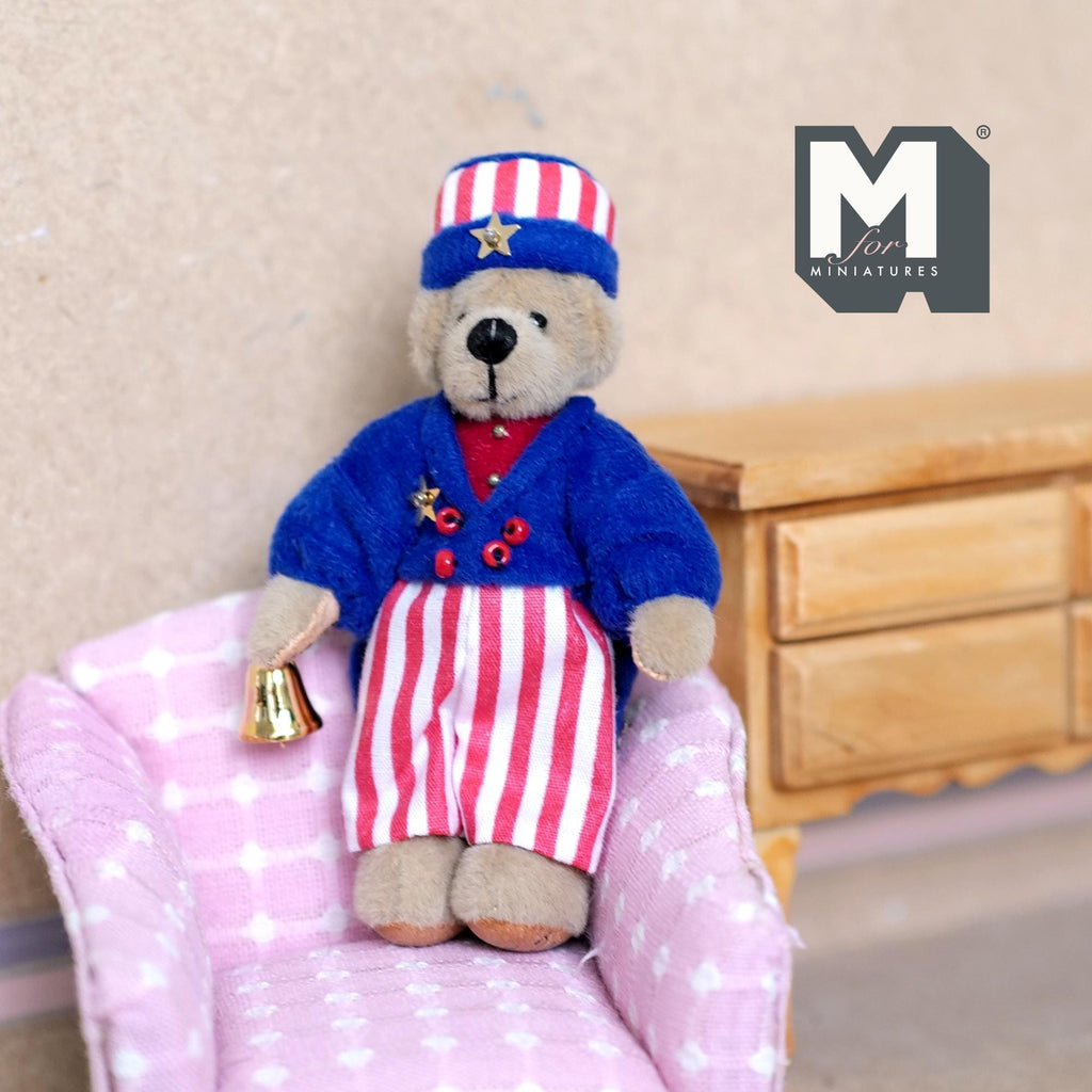 1:12 scale miniature plush bear in a stars and stripes outfit with cap and bell, shown sitting on a pink armchair