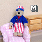 1:12 scale miniature plush bear in a stars and stripes outfit with cap and bell, shown sitting on a pink armchair