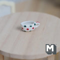 1:12 scale miniature ceramic cream boat with handle, white with red and green polka dots, shown on a round wood table
