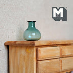1:12 scale miniature green glass bottle vase with round bulb shape and flared rim, translucent, on a wood dresser