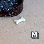 1:12 scale miniature dog bone treat in white, shown on a craft board beside a wicker pet bed
