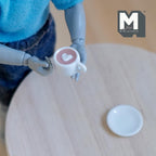 1:12 scale miniature hot chocolate cup with heart cream and white saucer, shown held by a poseable figure
